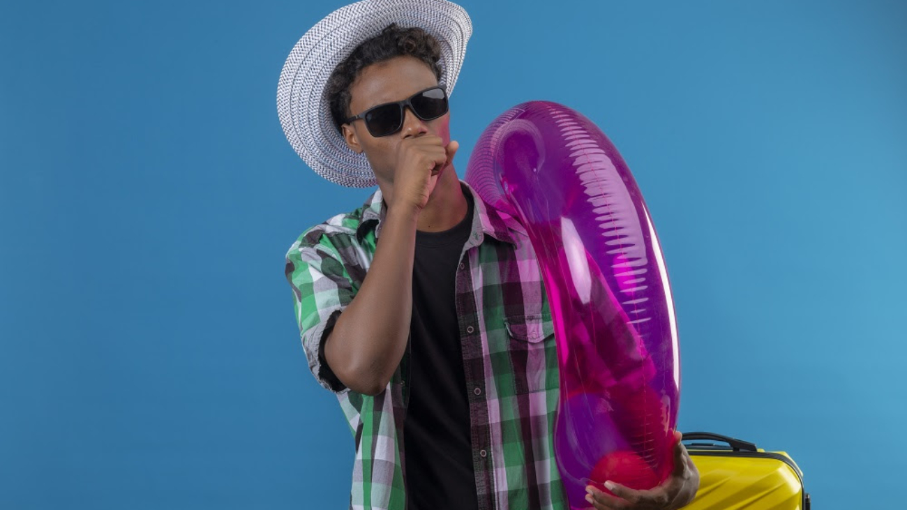 young african american traveler man in summer hat wearing black sunglasses standing with suitcase holding inflatable ring coughing over blue background